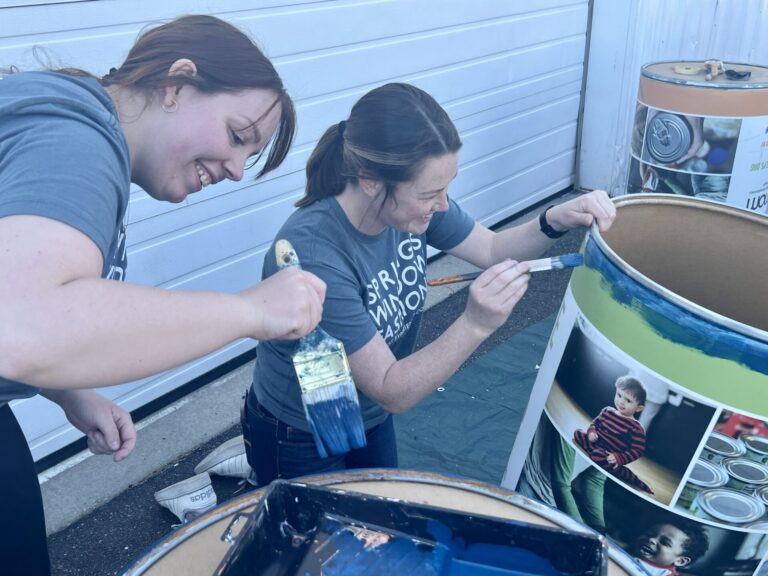 Two white women wearing matching gray T-shirts crouch in front of food donation barrels with paintbrushes.