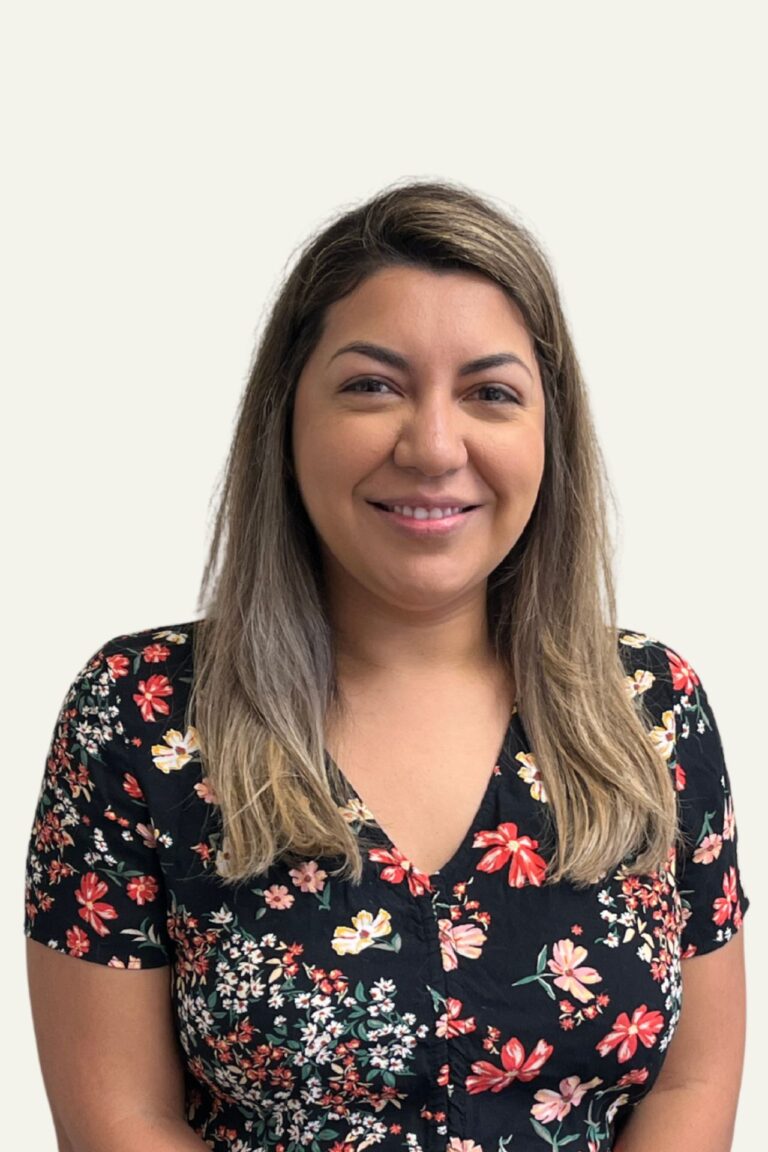 A headshot photo of Melissa Martinez smiling standing in front of a cream colored wall wearing a flower print blouse.