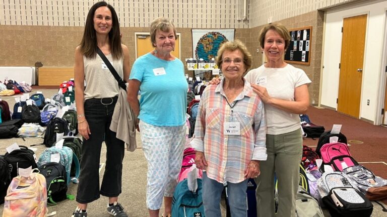 A group of female volunteers standing among donated backpacks and school supplies.